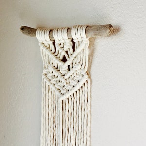 May include: A white macrame wall hanging with a wooden dowel at the top. The macrame is in a geometric pattern with fringe at the bottom.