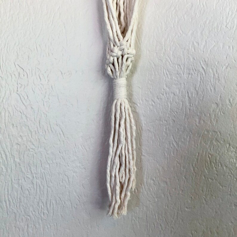 May include: A white macrame plant hanger with a tassel. The hanger is made of cotton cord and has a simple design.