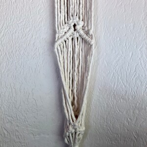 May include: A white macrame plant hanger with a knotted design. The hanger is made of thick cotton cord and has a triangular shape.