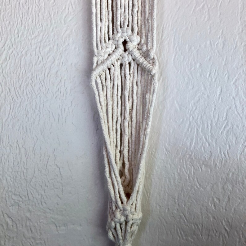 May include: A white macrame plant hanger with a knotted design. The hanger is made of thick cotton cord and has a triangular shape.