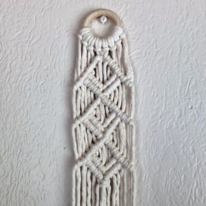 May include: A white macrame wall hanging with a wooden ring at the top. The macrame is made with a diamond pattern.