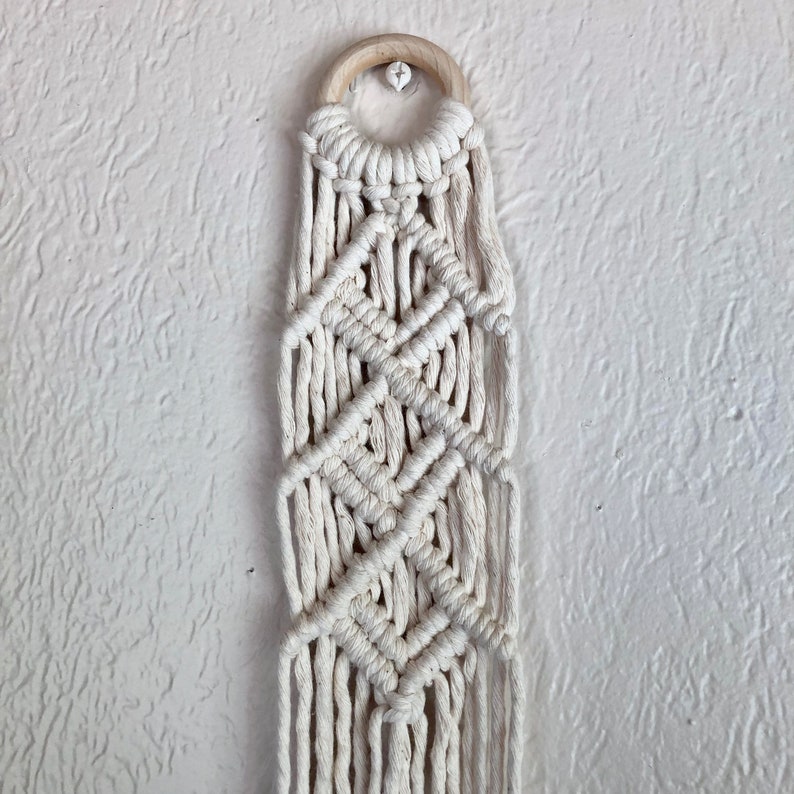 May include: A white macrame wall hanging with a wooden ring at the top. The macrame is made with a diamond pattern.