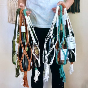 Peut inclure: Trois bouteilles de vin sont tenues dans des porte-bouteilles en macramé blanc, marron et bleu. Les bouteilles sont toutes de marques différentes et ont des étiquettes différentes.