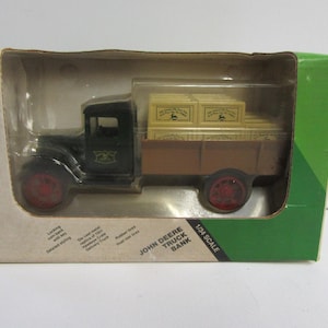 Peut inclure: Une boîte verte et jaune contenant une tirelire en forme de camion John Deere. Le camion est noir avec des roues rouges et transporte des caisses en bois. La boîte porte l'inscription "John Deere Truck Bank 1/24 Scale" sur le devant.