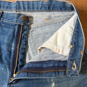 K&ouml;nnte beinhalten: Nahaufnahme einer blauen Jeans mit teilweise ge&ouml;ffnetem Messingrei&szlig;verschluss und Knopfverschluss. Die Jeans weist Gebrauchsspuren auf, mit wei&szlig;en Farbspritzern und einem hellen Innenfutter.