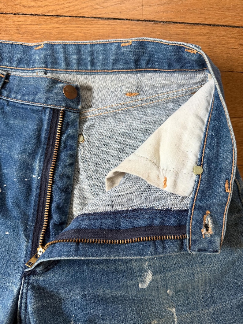 K&ouml;nnte beinhalten: Nahaufnahme einer blauen Jeans mit teilweise ge&ouml;ffnetem Messingrei&szlig;verschluss und Knopfverschluss. Die Jeans weist Gebrauchsspuren auf, mit wei&szlig;en Farbspritzern und einem hellen Innenfutter.