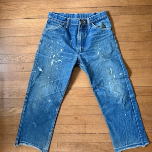 K&ouml;nnte beinhalten: Blaue Jeans mit wei&szlig;en Farbspritzern. Die Jeans haben ein klassisches F&uuml;nf-Taschen-Design, einen Knopf- und Rei&szlig;verschluss und einen geraden Beinabschluss. Die Jeans werden auf einer Holzoberfl&auml;che pr&auml;sentiert.