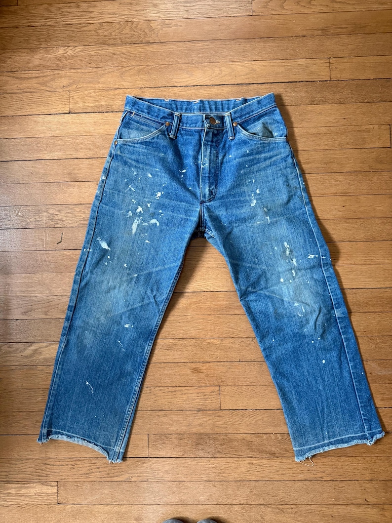 K&ouml;nnte beinhalten: Blaue Jeans mit wei&szlig;en Farbspritzern. Die Jeans haben ein klassisches F&uuml;nf-Taschen-Design, einen Knopf- und Rei&szlig;verschluss und einen geraden Beinabschluss. Die Jeans werden auf einer Holzoberfl&auml;che pr&auml;sentiert.