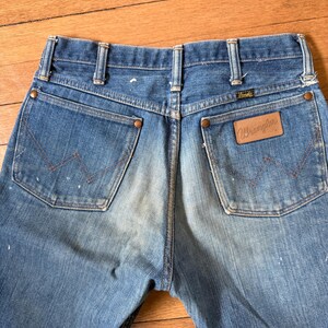 K&ouml;nnte beinhalten: Nahaufnahme der R&uuml;ckseite einer blauen Wrangler-Jeans. Die Jeans hat zwei Ges&auml;&szlig;taschen mit dem Wrangler-Logo auf der rechten Tasche. Die Jeans weist Gebrauchsspuren auf, mit einigen wei&szlig;en Farbspritzern.
