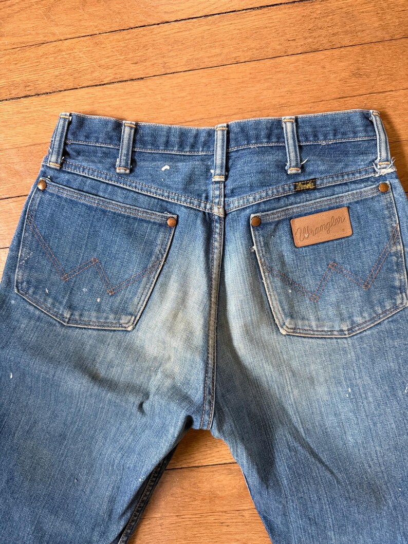 K&ouml;nnte beinhalten: Nahaufnahme der R&uuml;ckseite einer blauen Wrangler-Jeans. Die Jeans hat zwei Ges&auml;&szlig;taschen mit dem Wrangler-Logo auf der rechten Tasche. Die Jeans weist Gebrauchsspuren auf, mit einigen wei&szlig;en Farbspritzern.