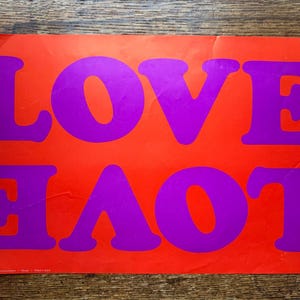 May include: A rectangular poster with the word "LOVE" in large, purple, bold letters against a bright orange background. The poster is slightly creased and has a vintage aesthetic.