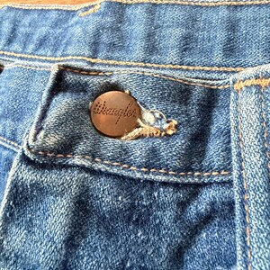 K&ouml;nnte beinhalten: Nahaufnahme einer blauen Jeans mit einem kupferfarbenen Knopf. Der Knopf ist mit dem Wort "Wrangler" gepr&auml;gt. Die Jeans hat sichtbare N&auml;hte und ein klassisches Design.