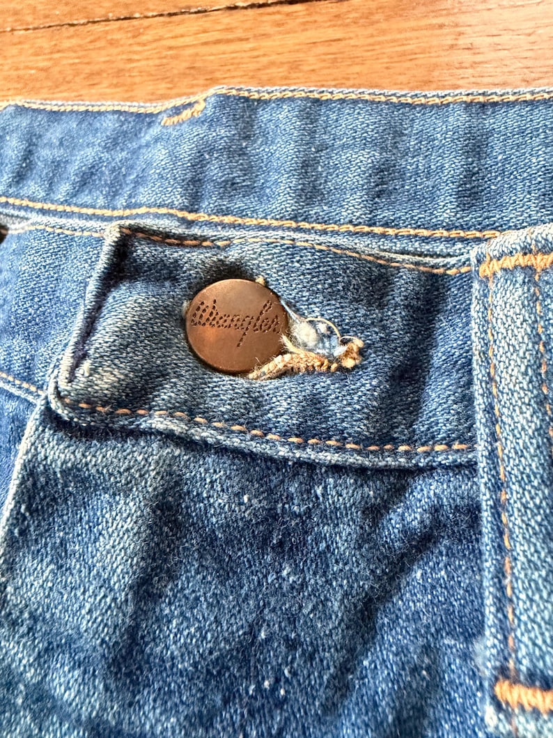 K&ouml;nnte beinhalten: Nahaufnahme einer blauen Jeans mit einem kupferfarbenen Knopf. Der Knopf ist mit dem Wort "Wrangler" gepr&auml;gt. Die Jeans hat sichtbare N&auml;hte und ein klassisches Design.