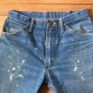 K&ouml;nnte beinhalten: Nahaufnahme einer blauen Jeans mit Farbspritzern. Die Jeans hat einen Knopfverschluss und ein klassisches F&uuml;nf-Taschen-Design. Der Denim ist verblichen und weist Gebrauchsspuren auf. Die Jeans ist ein klassischer Stil.