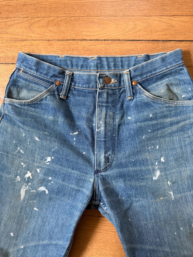 K&ouml;nnte beinhalten: Nahaufnahme einer blauen Jeans mit Farbspritzern. Die Jeans hat einen Knopfverschluss und ein klassisches F&uuml;nf-Taschen-Design. Der Denim ist verblichen und weist Gebrauchsspuren auf. Die Jeans ist ein klassischer Stil.