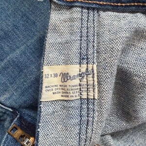 K&ouml;nnte beinhalten: Nahaufnahme einer blauen Jeans aus Denim mit einem Messingrei&szlig;verschluss und einem "Wrangler"-Etikett. Das Etikett enth&auml;lt Waschanweisungen und die Gr&ouml;&szlig;e 32x30. Hergestellt in den USA.