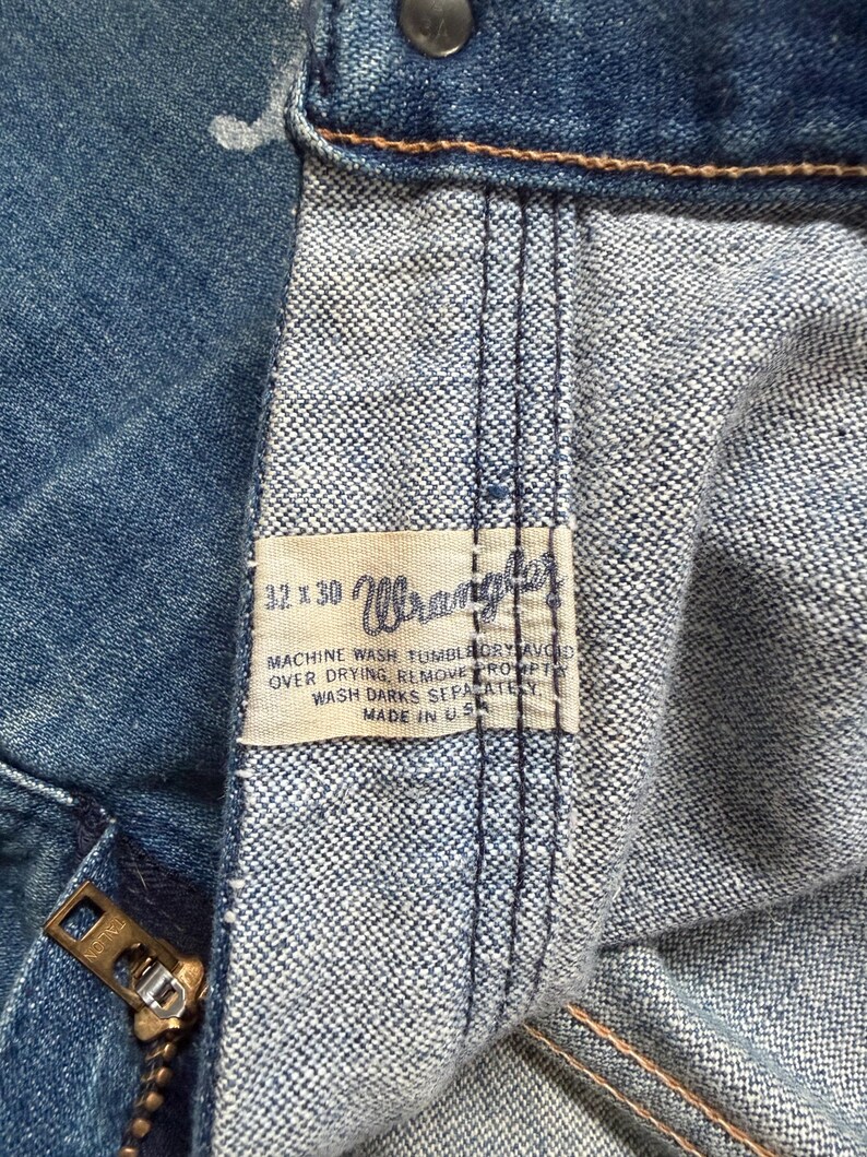 K&ouml;nnte beinhalten: Nahaufnahme einer blauen Jeans aus Denim mit einem Messingrei&szlig;verschluss und einem "Wrangler"-Etikett. Das Etikett enth&auml;lt Waschanweisungen und die Gr&ouml;&szlig;e 32x30. Hergestellt in den USA.
