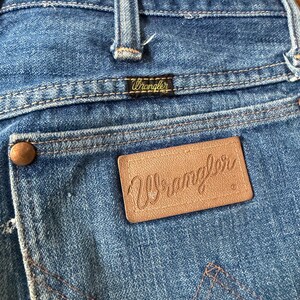 K&ouml;nnte beinhalten: Nahaufnahme der R&uuml;ckseite einer blauen Jeans. Der Markenname Wrangler ist auf einem kleinen schwarzen Etikett und einem gr&ouml;&szlig;eren braunen Lederaufn&auml;her zu sehen. Die Jeans hat klassische N&auml;hte und einen kupferfarbenen Knopf.