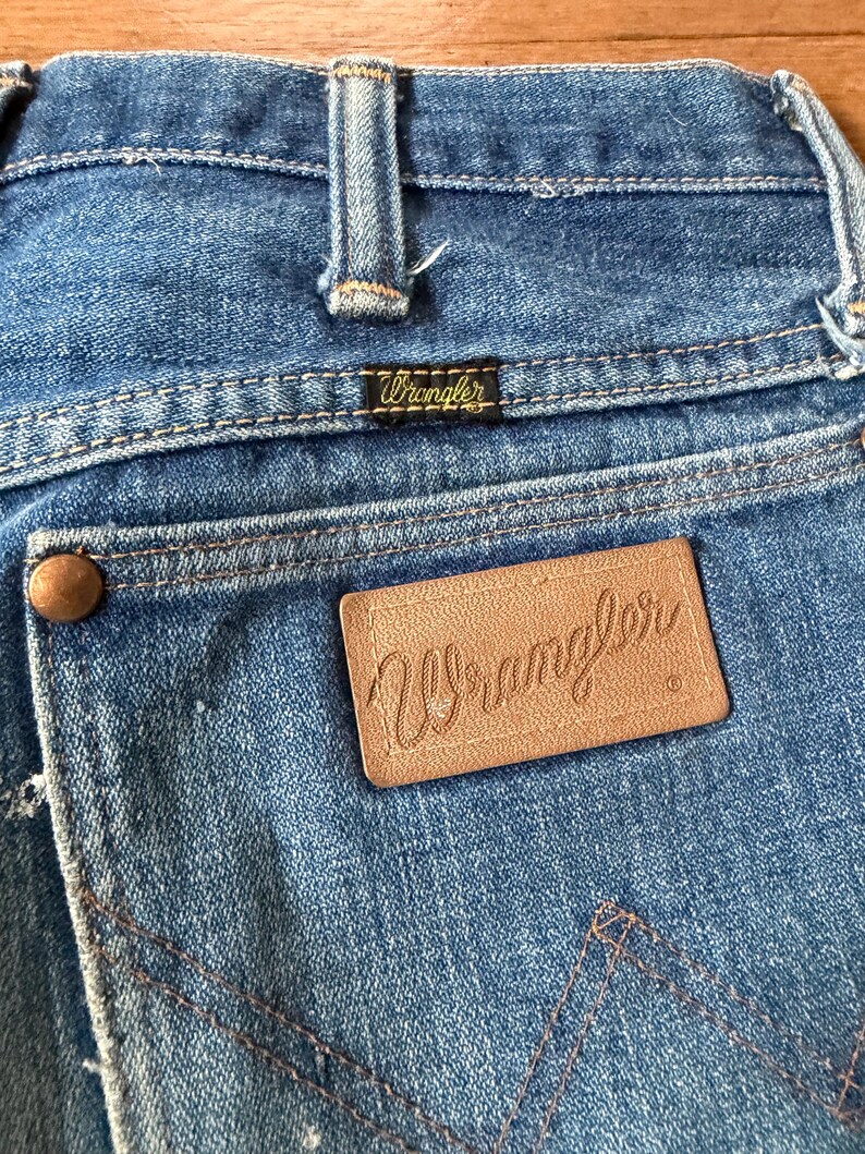 K&ouml;nnte beinhalten: Nahaufnahme der R&uuml;ckseite einer blauen Jeans. Der Markenname Wrangler ist auf einem kleinen schwarzen Etikett und einem gr&ouml;&szlig;eren braunen Lederaufn&auml;her zu sehen. Die Jeans hat klassische N&auml;hte und einen kupferfarbenen Knopf.