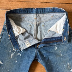 K&ouml;nnte beinhalten: Nahaufnahme einer blauen Jeans mit teilweise ge&ouml;ffnetem Rei&szlig;verschluss. Die Jeans hat wei&szlig;e Farbspritzer und sichtbare N&auml;hte. Das Innenfutter ist hellblau.