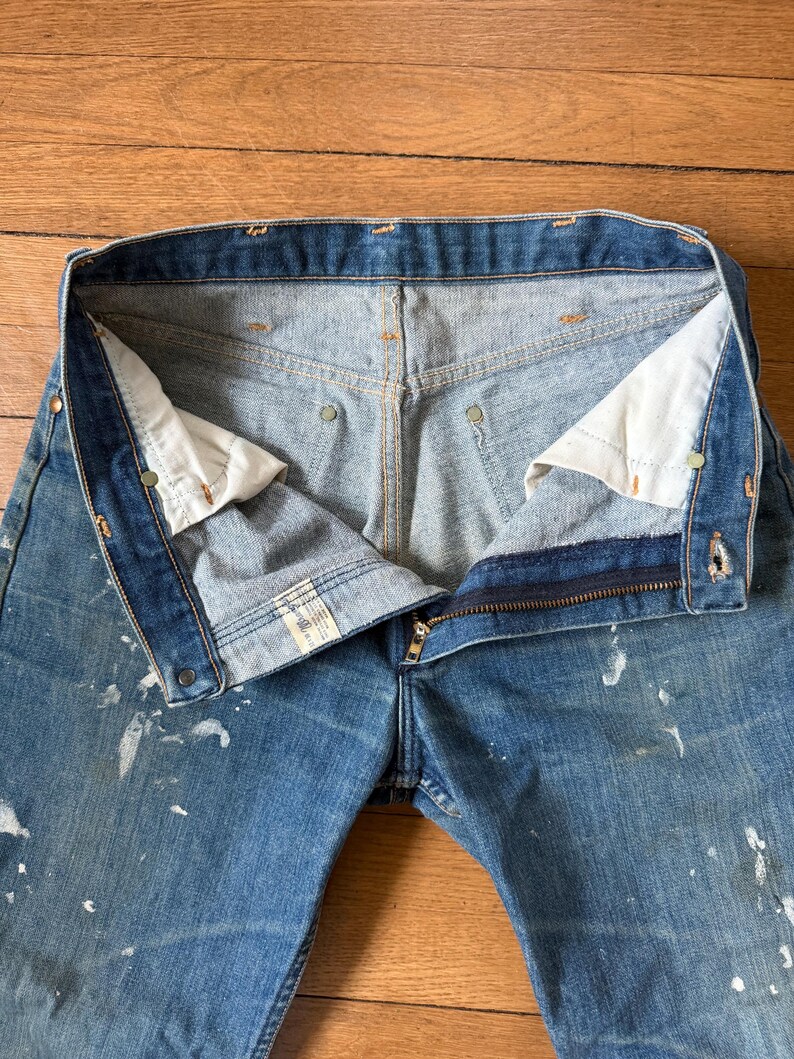 K&ouml;nnte beinhalten: Nahaufnahme einer blauen Jeans mit teilweise ge&ouml;ffnetem Rei&szlig;verschluss. Die Jeans hat wei&szlig;e Farbspritzer und sichtbare N&auml;hte. Das Innenfutter ist hellblau.