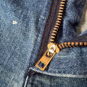 K&ouml;nnte beinhalten: Nahaufnahme eines blauen Jeanskleidungsst&uuml;cks mit einem Messingrei&szlig;verschluss. Der Rei&szlig;verschluss ist teilweise ge&ouml;ffnet und zeigt die Z&auml;hne und den Markennamen Talon auf dem Zipper. Der Denim hat ein abgenutztes Aussehen.