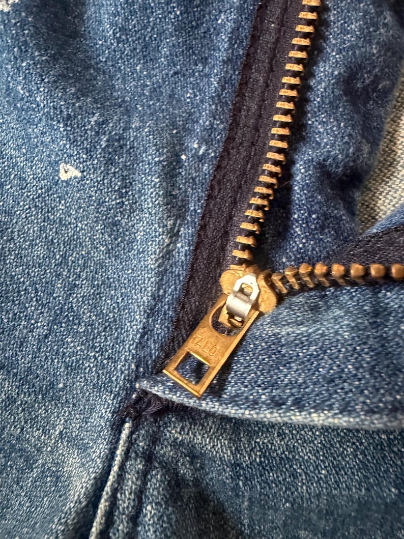 K&ouml;nnte beinhalten: Nahaufnahme eines blauen Jeanskleidungsst&uuml;cks mit einem Messingrei&szlig;verschluss. Der Rei&szlig;verschluss ist teilweise ge&ouml;ffnet und zeigt die Z&auml;hne und den Markennamen Talon auf dem Zipper. Der Denim hat ein abgenutztes Aussehen.