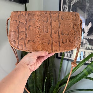 May include: A brown snakeskin-patterned shoulder bag with a curved flap and braided strap. The bag is held up by a hand, showcasing the texture and color variations of the material. The bag's design is vintage and classic.
