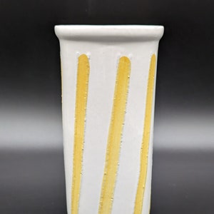 May include: A white ceramic vase with vertical yellow stripes. The vase has a cylindrical shape with a slightly flared top. The yellow stripes are evenly spaced and add a decorative element to the vase.