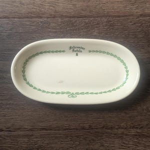 May include: An oval, off-white ceramic serving platter with a green leaf border and the text "Schroeder Hotels" in green. The platter is empty and sits on a dark wood surface.