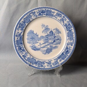 May include: A white ceramic plate with a blue floral border and a central landscape scene. The plate features a detailed design of a building, trees, and figures, creating a classic aesthetic. The plate is displayed on a clear plastic stand.