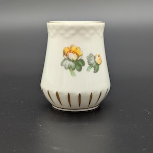 May include: A white ceramic salt shaker with a floral design. The shaker features a gold rim and base accents. The floral design includes yellow and orange flowers with green leaves. The top of the shaker has a textured pattern.