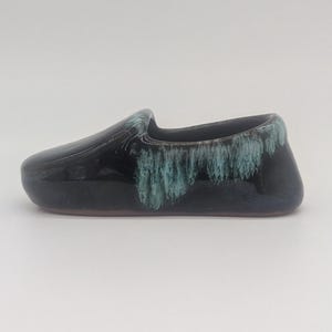 May include: A ceramic shoe-shaped vessel with a glossy dark navy blue finish. The upper edge features a decorative drip glaze effect in a light teal color that cascades down the sides of the object, creating a textured appearance against the smooth base.