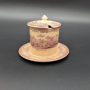 May include: A vintage ceramic sugar bowl with a lid and saucer. The off-white bowl and saucer feature a repeating floral pattern and a detailed landscape scene in a reddish-purple hue. The lid has a small knob and a slot for a spoon.