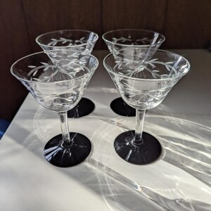 May include: Four clear glass cocktail glasses with etched floral designs. The glasses have a classic coupe shape with a clear stem and a black base. The glasses are arranged on a white surface.