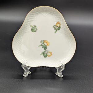 May include: White porcelain serving dish with a pear-like shape, featuring a textured surface and gold trim. The dish is adorned with hand-painted floral designs in yellow, green, and white. It is displayed on a clear, decorative stand.
