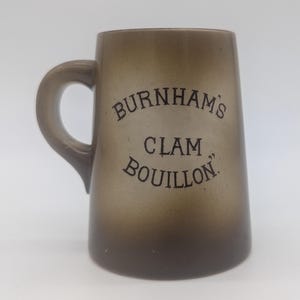 May include: A brown, tapered ceramic mug with a single handle. The front features dark brown text that reads "BURNHAM'S CLAM BOUILLON." The mug has a smooth, gradient finish that transitions from a lighter brown at the top to a darker brown at the base.