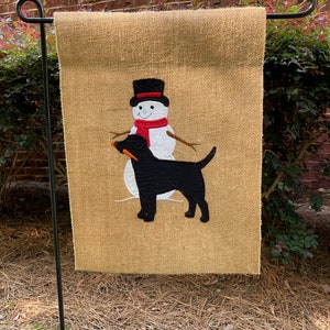 May include: A burlap garden flag featuring a snowman with a black top hat and a red scarf, and a black dog holding a carrot. The flag is displayed on a black metal stand, perfect for outdoor decor.