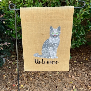 May include: A burlap welcome sign with a gray cat with a white pattern and green eyes. The sign says "Welcome" in black text.