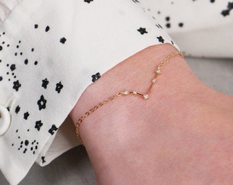 May include: A delicate gold-colored bracelet with small, sparkling stones is worn on a wrist. The bracelet has a V-shaped design with a series of small, round stones. The background includes a white blouse with a black star pattern.