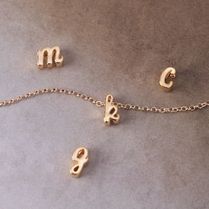 May include: A gold-colored necklace with the letter "k" as a pendant, along with the letters "m", "c", and "g" scattered around. The necklace and letters are on a neutral-toned background.