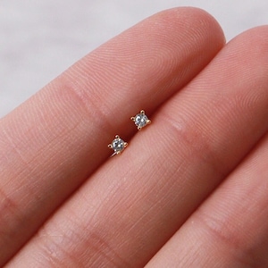 May include: A pair of small, square-shaped stud earrings with a sparkling, clear gemstone set in a gold-colored metal. The earrings are displayed on a person's finger, showcasing their delicate design and size.