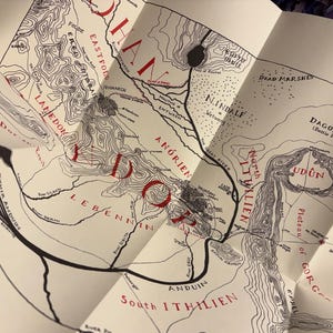 May include: A detailed map with black line art and red text, featuring geographical features and place names. The map is folded, revealing sections of a larger design. The map includes the text "Lamedon", "Udun", and "Daglorad".