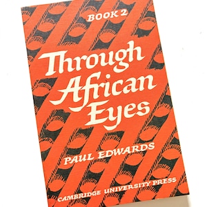 Pu&ograve; includere: Un libro vintage intitolato "Through African Eyes" di Paul Edwards. La copertina &egrave; arancione con un motivo diagonale nero e testo bianco. Il libro &egrave; etichettato "Book 2" e pubblicato da Cambridge University Press.