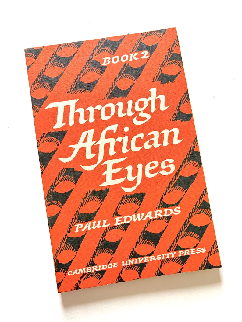 Pu&ograve; includere: Un libro vintage intitolato "Through African Eyes" di Paul Edwards. La copertina &egrave; arancione con un motivo diagonale nero e testo bianco. Il libro &egrave; etichettato "Book 2" e pubblicato da Cambridge University Press.