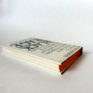 K&ouml;nnte beinhalten: Ein Buch mit dem Titel "Black Rage" mit schwarzem Text auf einem wei&szlig;en Einband. Der Buchr&uuml;cken ist schwarz und die Seiten sind cremefarben mit einem roten Rand. Das Buch ist leicht angewinkelt.