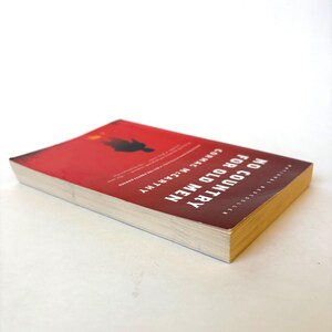May include: A paperback book titled "No Country for Old Men" by Cormac McCarthy. The cover is red with white text and a silhouette. The book's pages are off-white, and the edges are yellow.