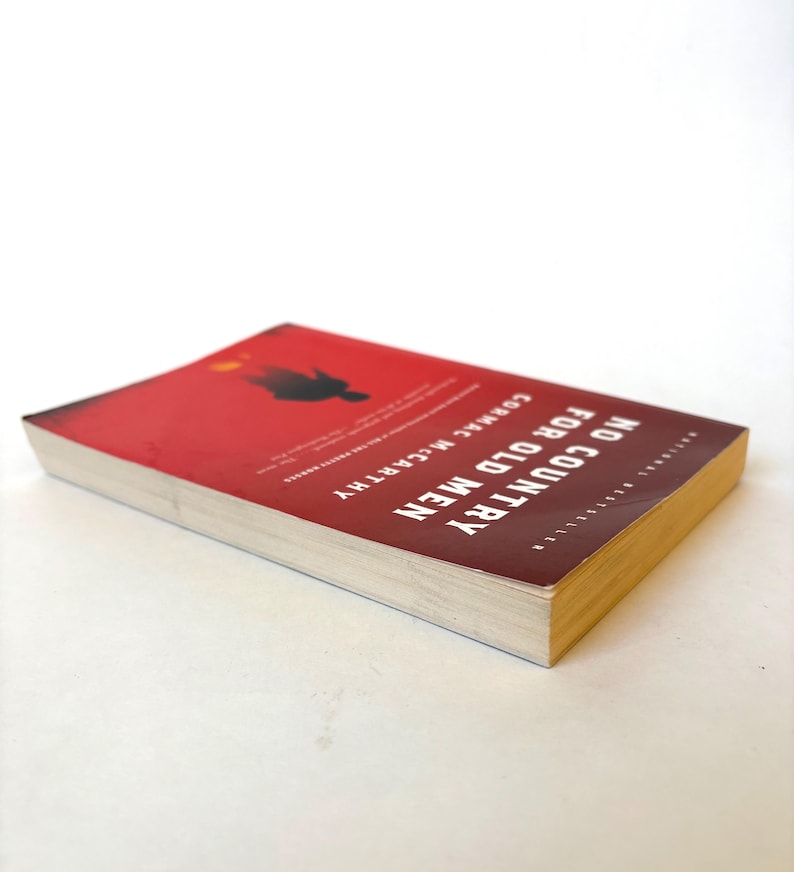 May include: A paperback book titled "No Country for Old Men" by Cormac McCarthy. The cover is red with white text and a silhouette. The book's pages are off-white, and the edges are yellow.