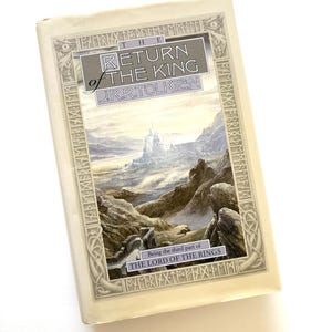 May include: A hardcover book titled "The Return of the King" by J.R.R. Tolkien, part of "The Lord of the Rings" series. The cover features a landscape illustration with a castle and mountains, framed by an ornate border. The book is in a protective cover.