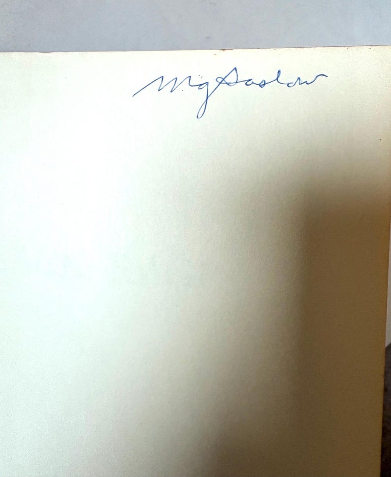 K&ouml;nnte beinhalten: Nahaufnahme eines cremefarbenen Papiers mit der handschriftlichen Signatur "mg Sadler" in blauer Tinte oben. Das Papier hat eine leicht strukturierte Oberfl&auml;che.